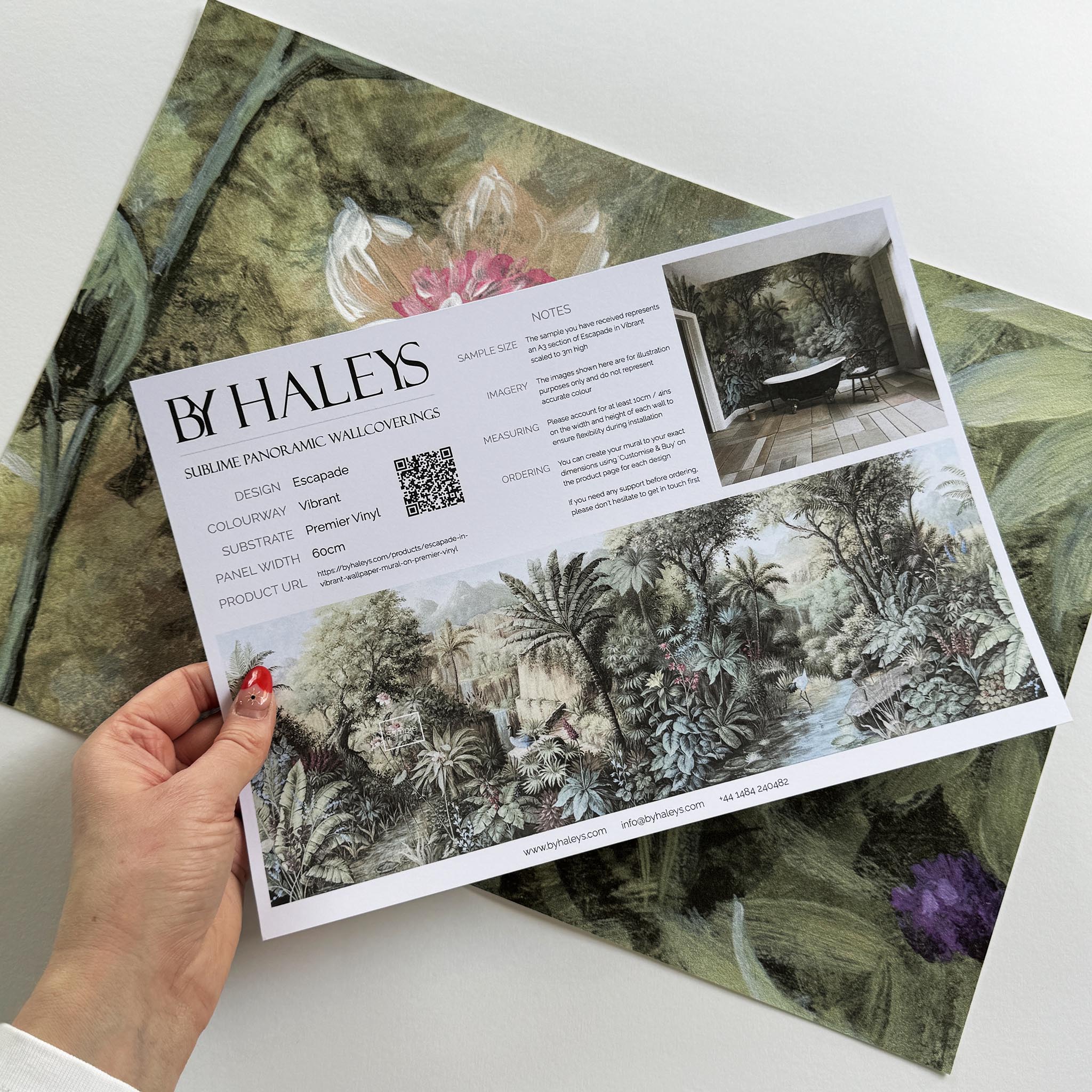 A hand showing information for a vinyl wallpaper mural sample which features a tropical paradise in cool greens and blushes.