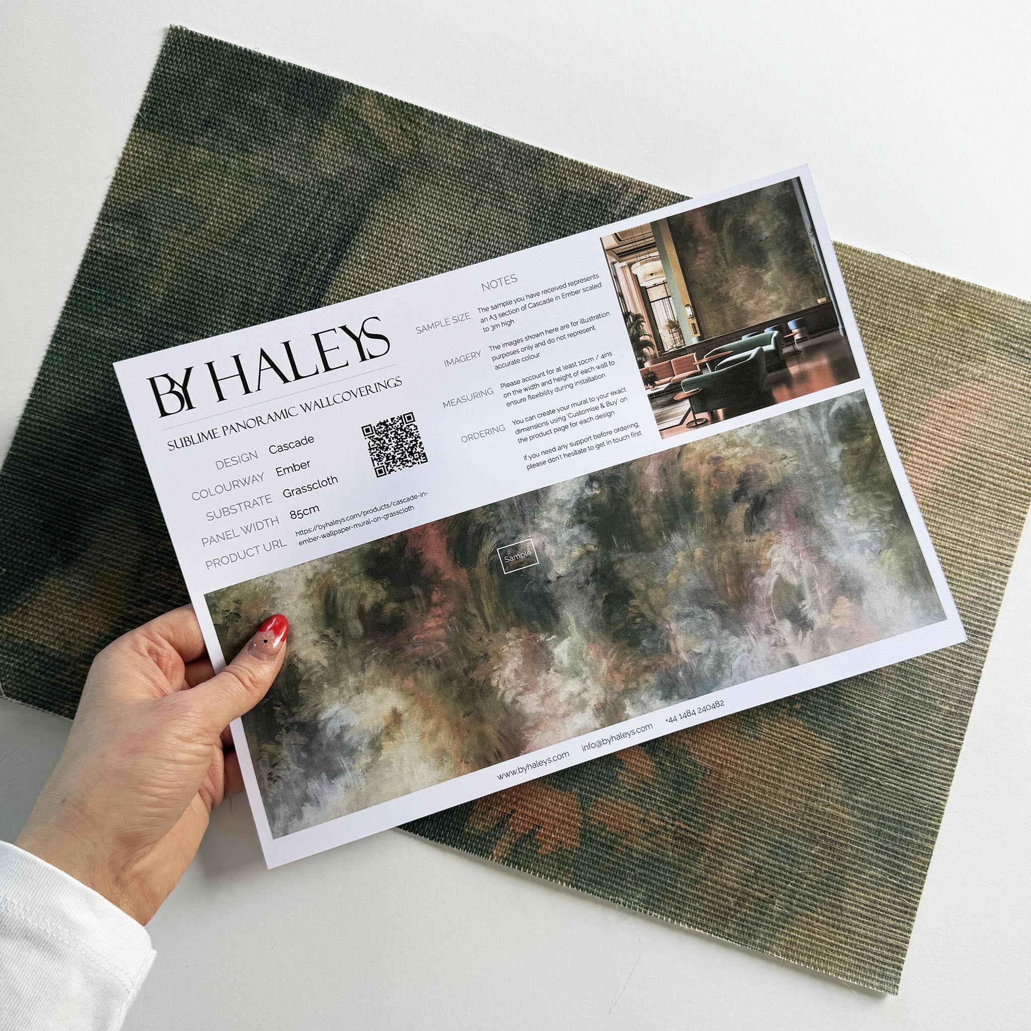 A hand showing information for a grasscloth wallpaper mural sample which features an abstract haze in green and blush.
