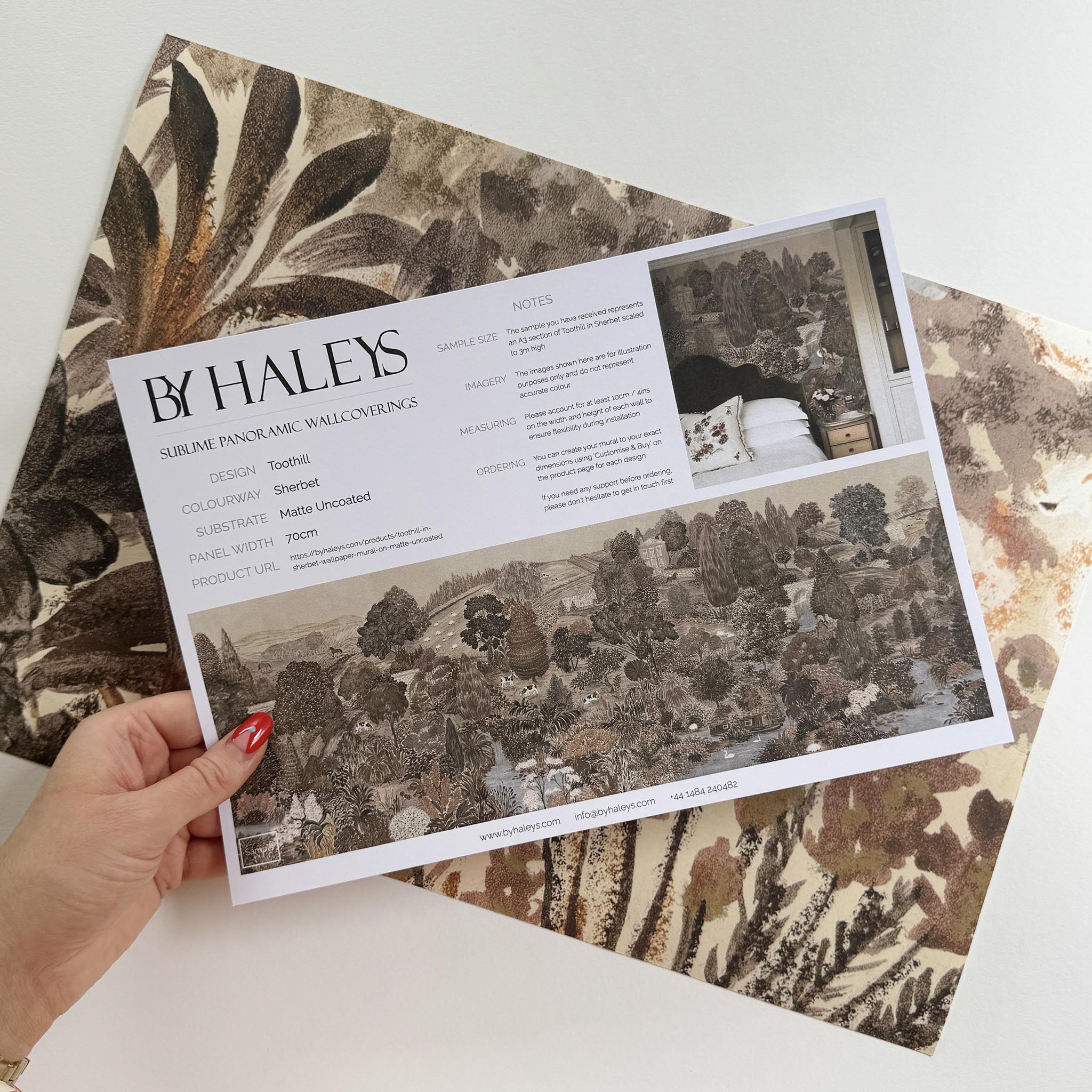 A hand showing information for a matte wallpaper mural sample which features a countryside in clay and soft plum tones