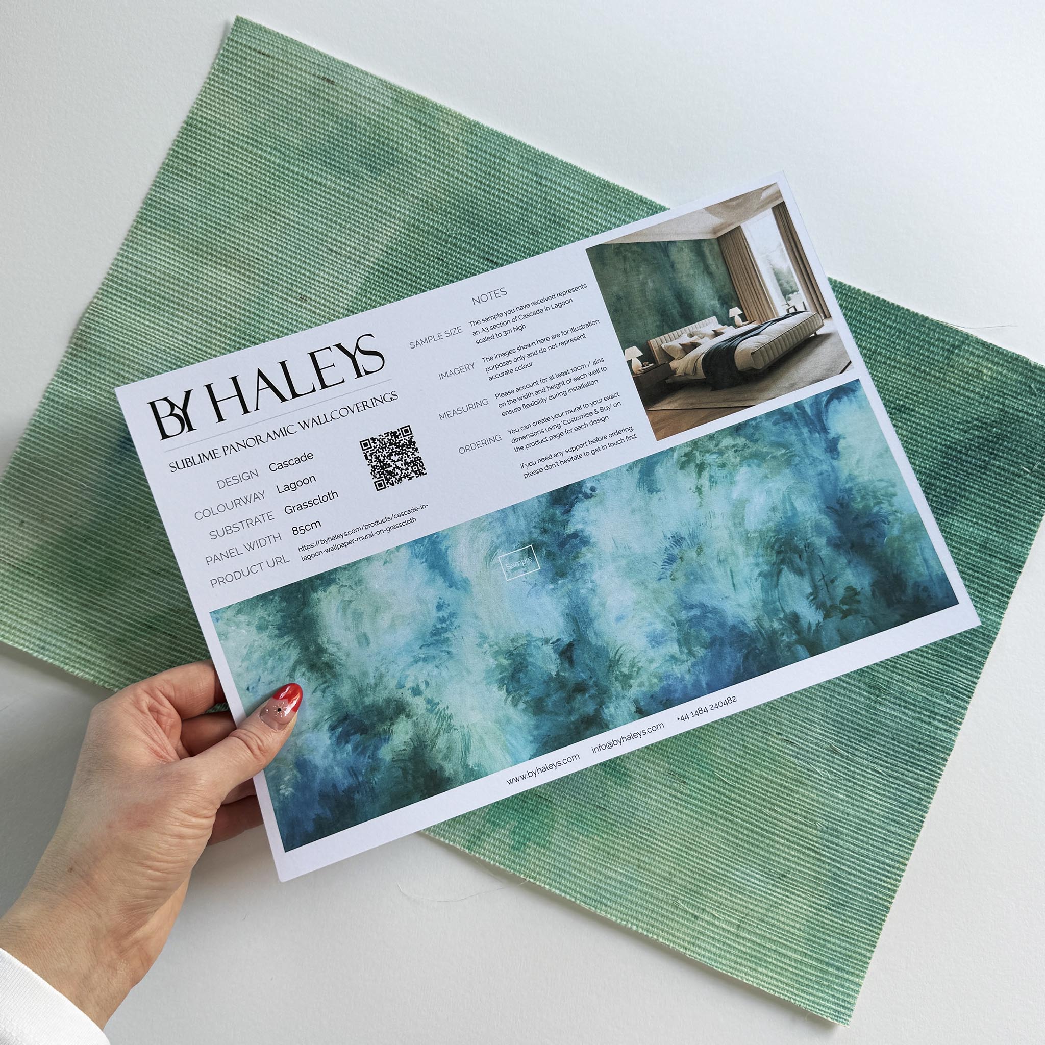 A hand showing information for a grasscloth wallpaper mural sample which features an abstract wash in aqua and jade.