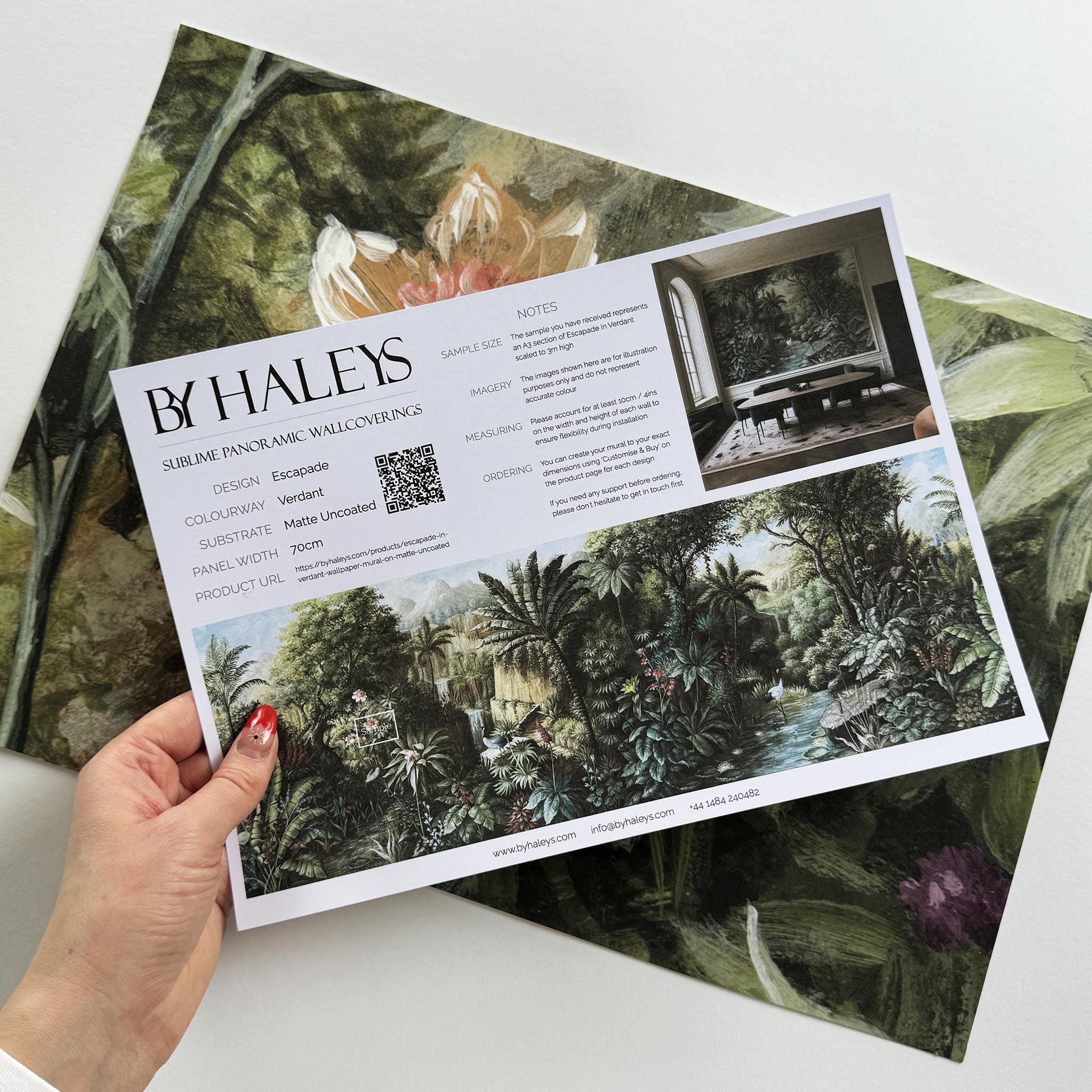A hand showing information for a matte wallpaper mural sample which features a tropical scene in green and pink.