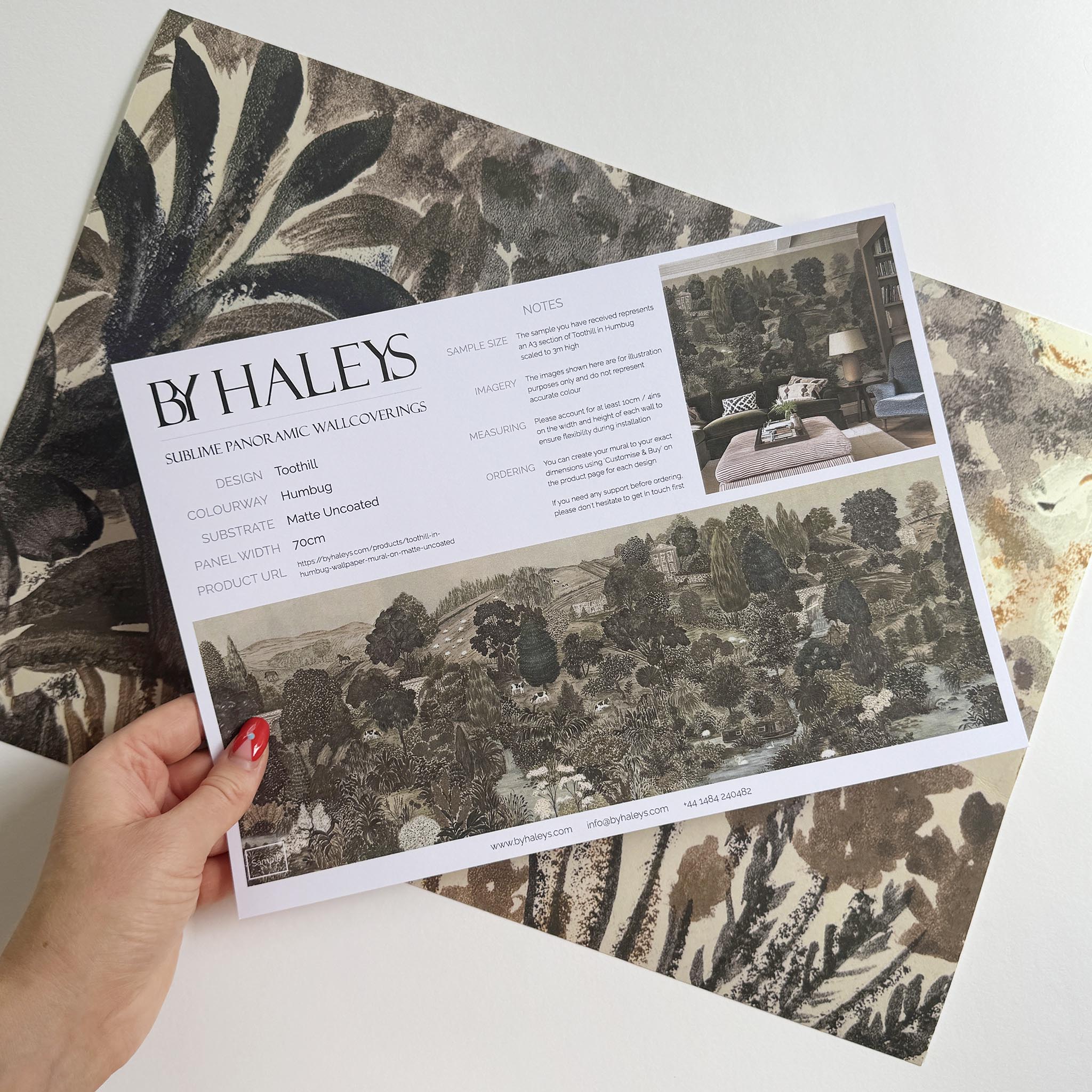 A hand showing information for a matte wallpaper mural sample which features a countryside in deep olive, slate and ash