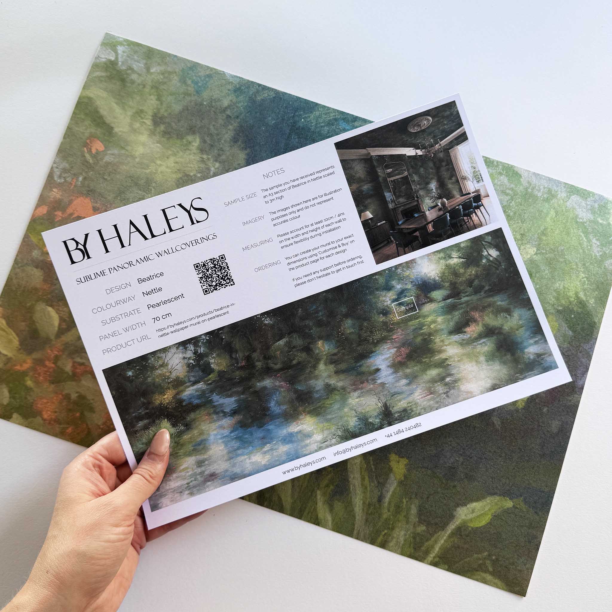 A hand showing information for a pearlescent wallpaper mural sample which features moody mural in nettle green, teal, ochre foliage.