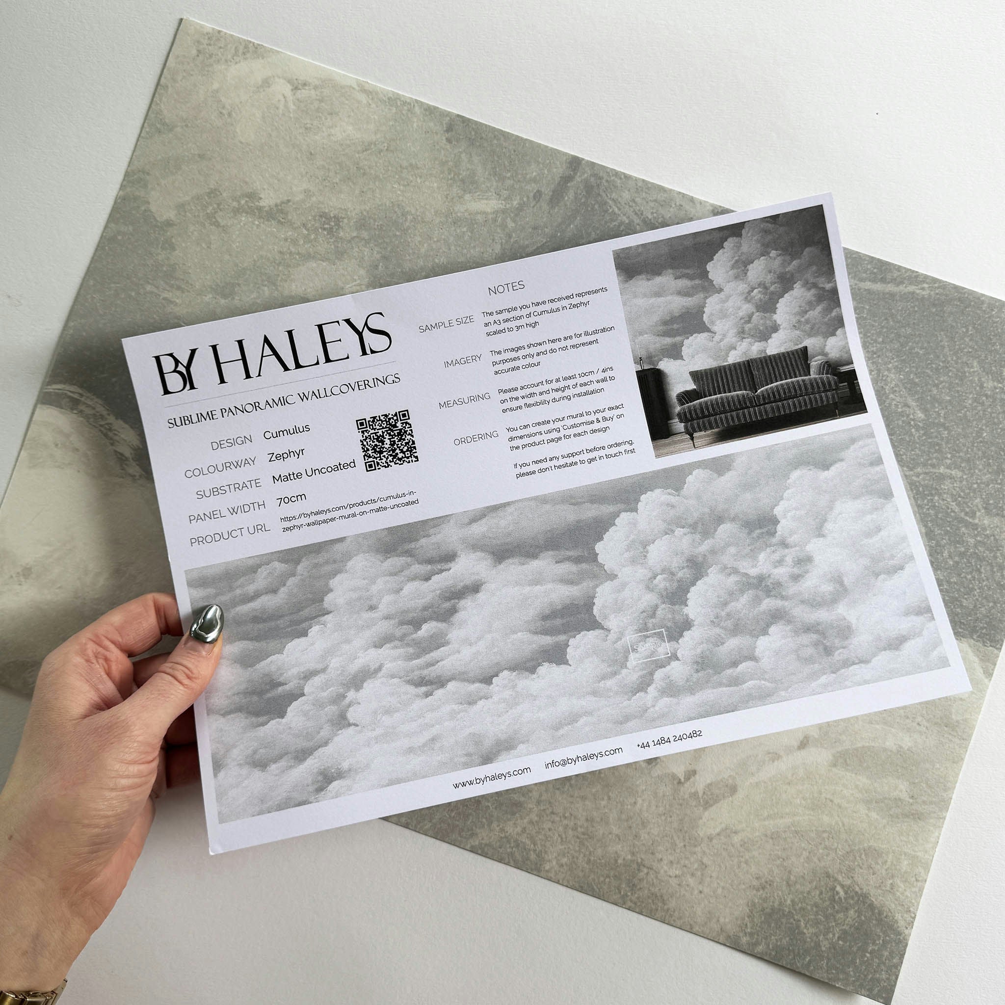 A hand showing information for a matte wallpaper mural sample which features pale sculpted clouds
