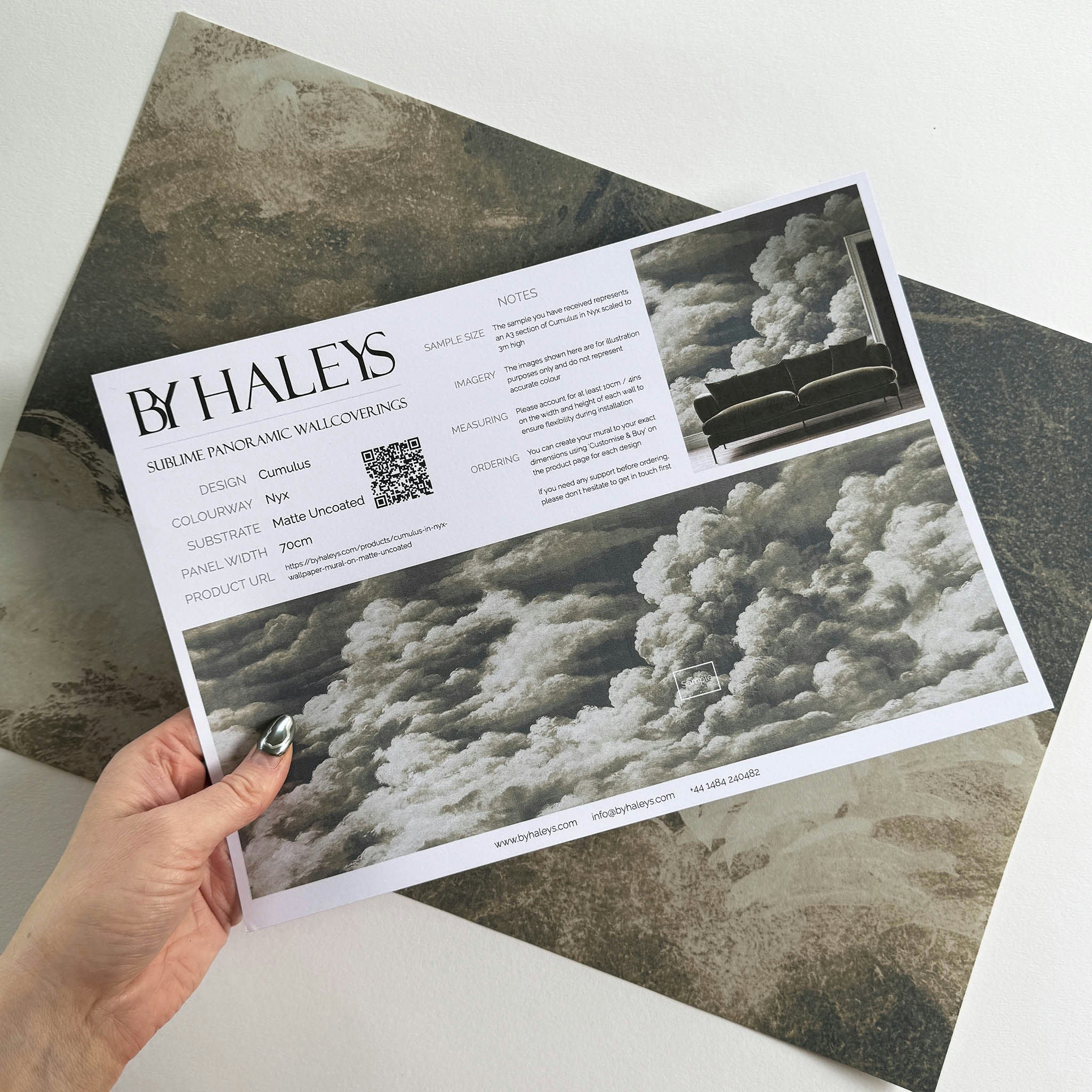 A hand showing information for a matte wallpaper mural sample which features dramatic cream clouds