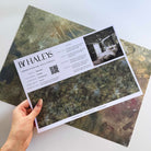 A hand showing information for a pearlescent wallpaper mural sample which features misty mural in fennel, sage, olive, indigo and ochre.