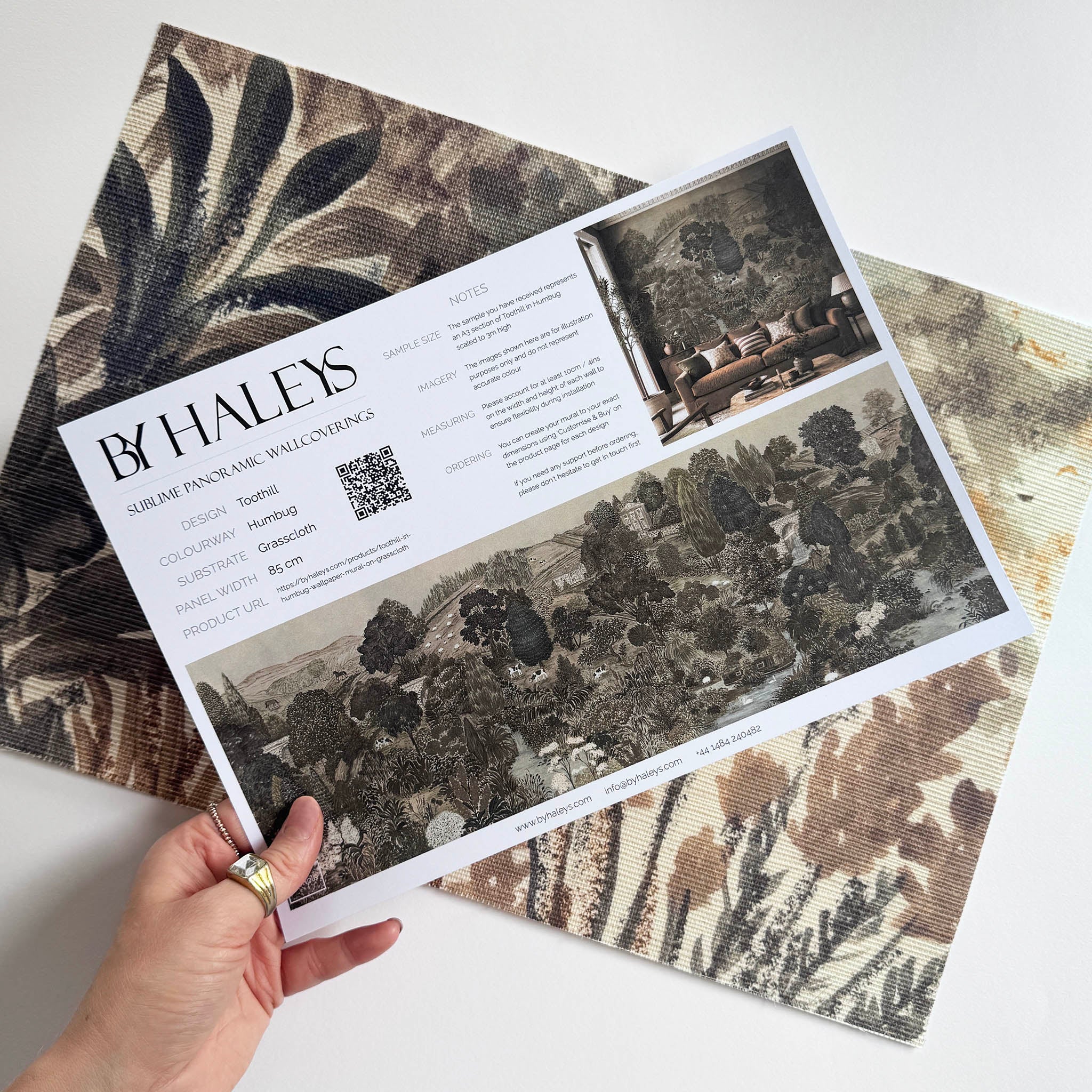 A hand showing information for a grasscloth wallpaper mural sample which features a countryside in teal, sea green and slate 