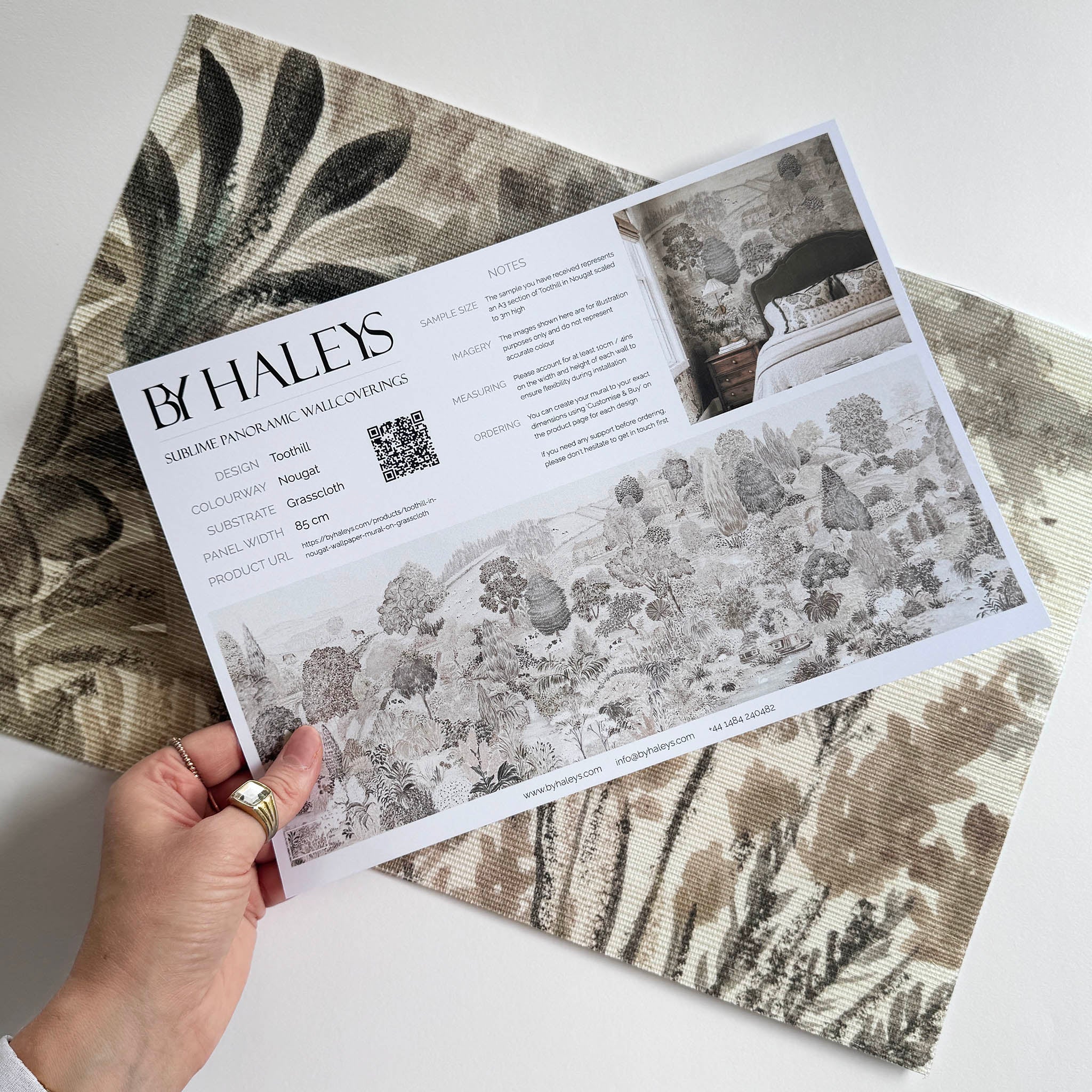 A hand showing information for a grasscloth wallpaper mural sample which features a countryside in teal, sea green and slate 