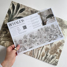 A hand showing information for a grasscloth wallpaper mural sample which features a countryside in teal, sea green and slate 